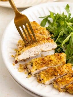 A fork picking up a slice of golden, breadcrumb-coated chicken breast next to a serving of fresh arugula on a white plate.