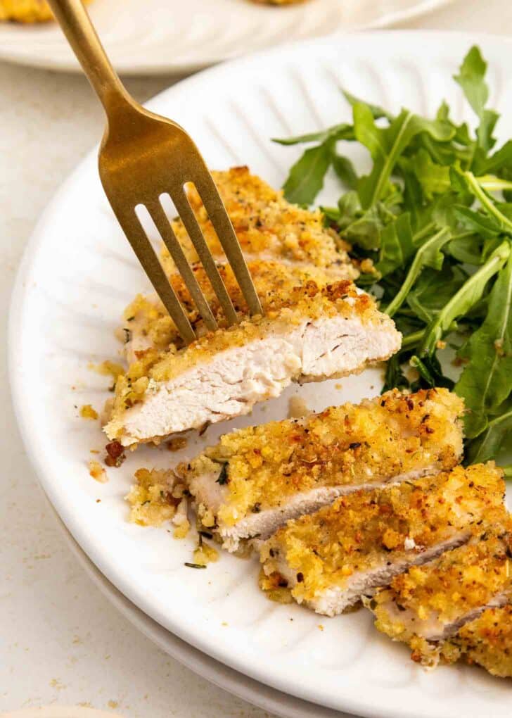 A fork picking up a slice of golden, breadcrumb-coated chicken breast next to a serving of fresh arugula on a white plate.