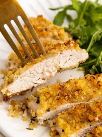 A close-up of a breaded and baked chicken cutlet sliced on a white plate, with a fork holding one piece. Fresh green arugula is served on the side.