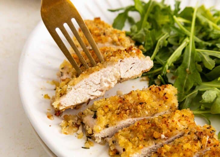 A close-up of a breaded and baked chicken cutlet sliced on a white plate, with a fork holding one piece. Fresh green arugula is served on the side.