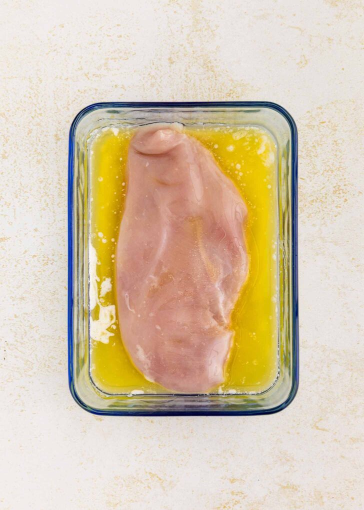 A raw chicken breast sits in a rectangular glass dish filled with yellow liquid on a light-colored surface.