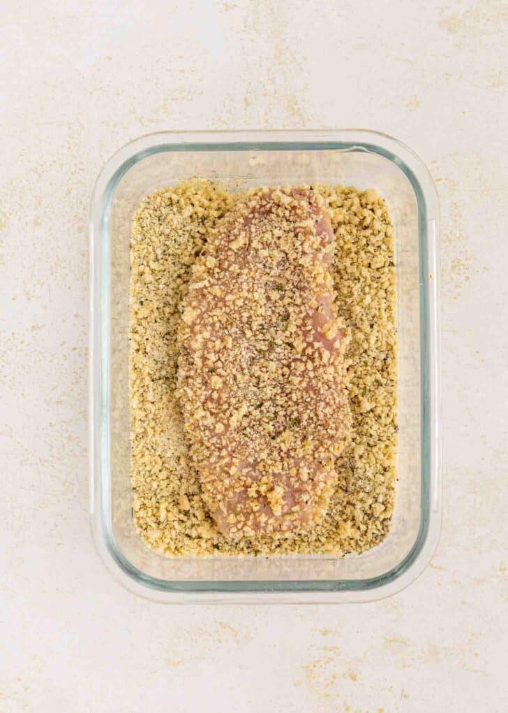 A raw chicken breast coated in breadcrumbs sits in a rectangular glass dish on a light-colored surface.