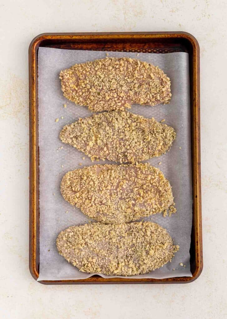 Four breaded cutlets are arranged in a single layer on a parchment-lined baking sheet, ready to be baked. The cutlets are evenly coated with a crumb mixture.