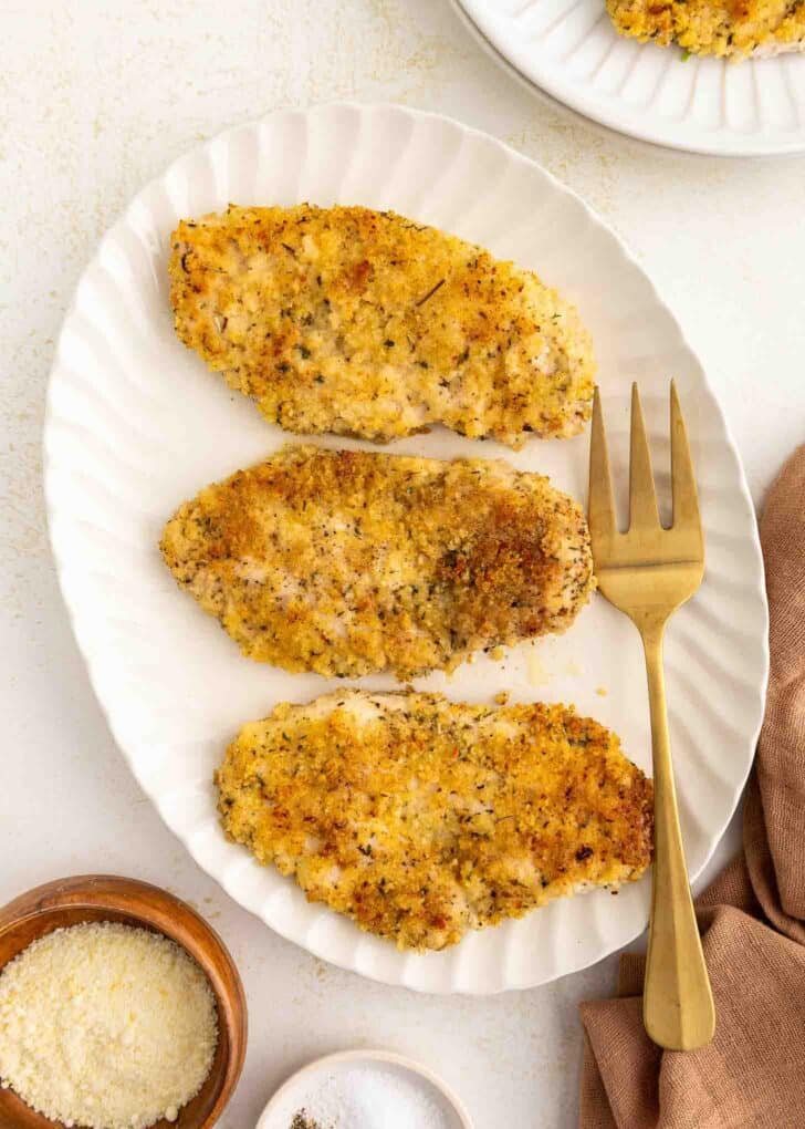 Three breaded and baked chicken cutlets are arranged on a white oval plate with a gold fork beside them. A small bowl of grated cheese and a bowl of salt are nearby on the table.