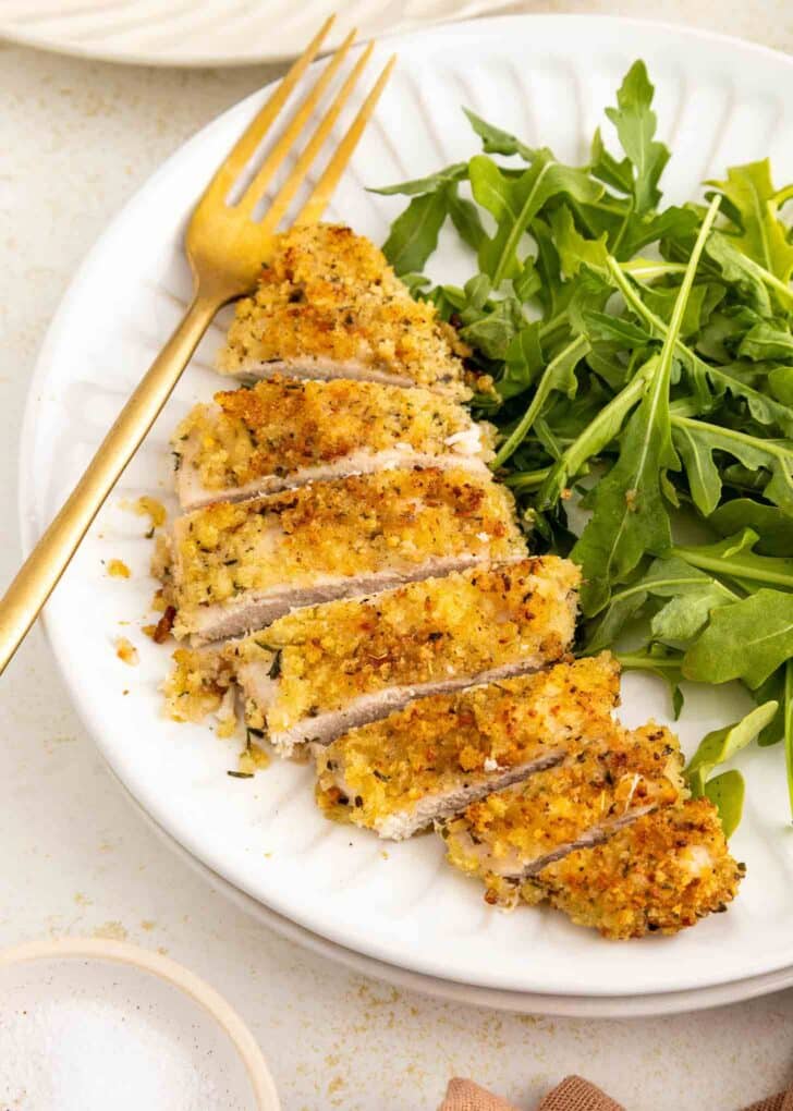 A white plate with sliced, breaded baked chicken breast and a serving of fresh arugula; a gold fork rests on the plate.