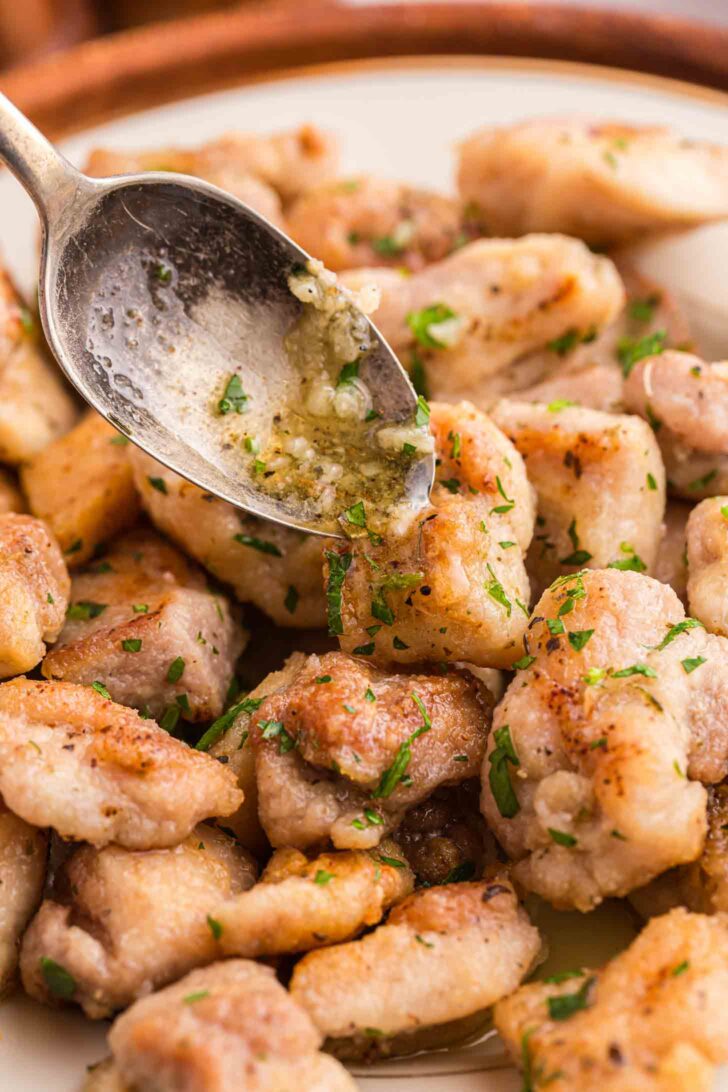 A spoon drizzles garlic butter sauce over golden, pan-seared pieces of chicken garnished with chopped parsley on a white plate.