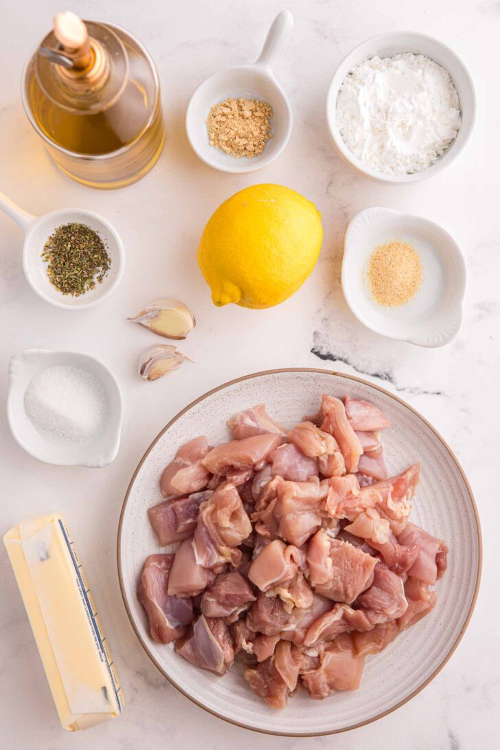 A plate of raw, cubed chicken surrounded by a lemon, garlic cloves, butter, a bottle of oil, and small bowls of seasonings, spices, and flour on a white marble surface.