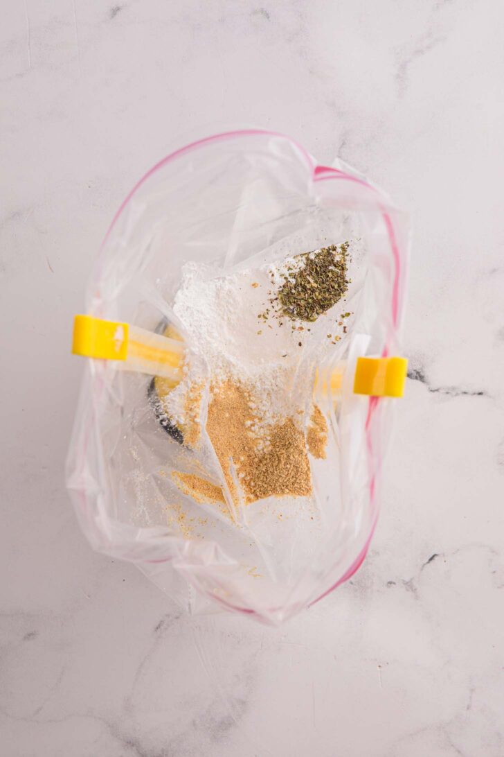 A plastic zip-top bag held open with clips contains various spices and seasonings, including brown and green powders, on a light marble surface.