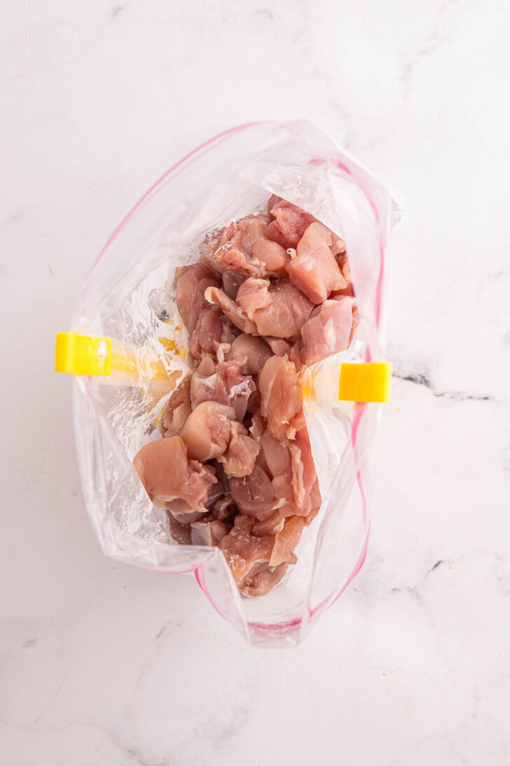 Raw, diced chicken placed inside a clear plastic zip-top bag that is held open with yellow bag holders, set on a white marble surface.