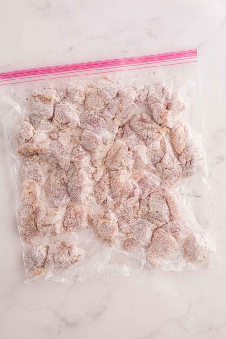 A clear ziplock bag filled with uncooked, flour-coated chicken pieces arranged in a single layer on a white marble surface.