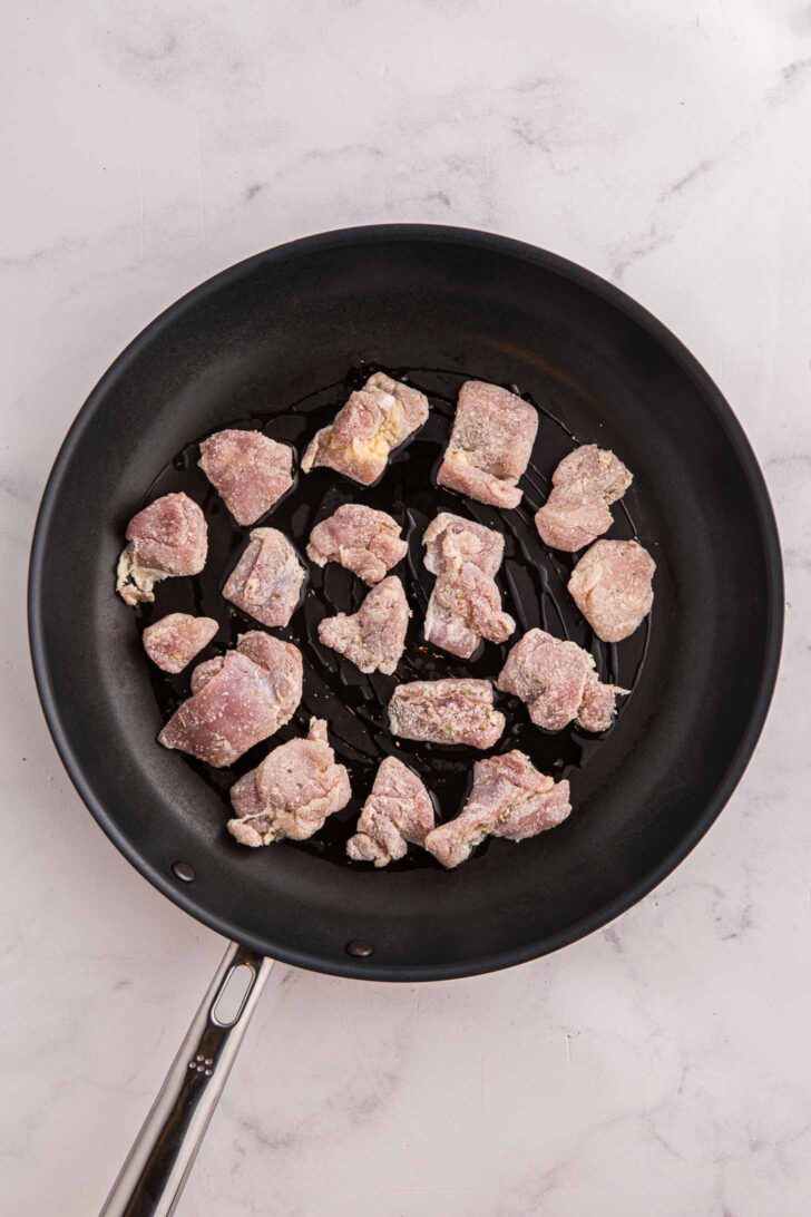 Raw, seasoned pieces of chicken are spread out in a single layer in a black nonstick frying pan with a silver handle, ready to be cooked. The pan is placed on a light-colored, marbled surface.