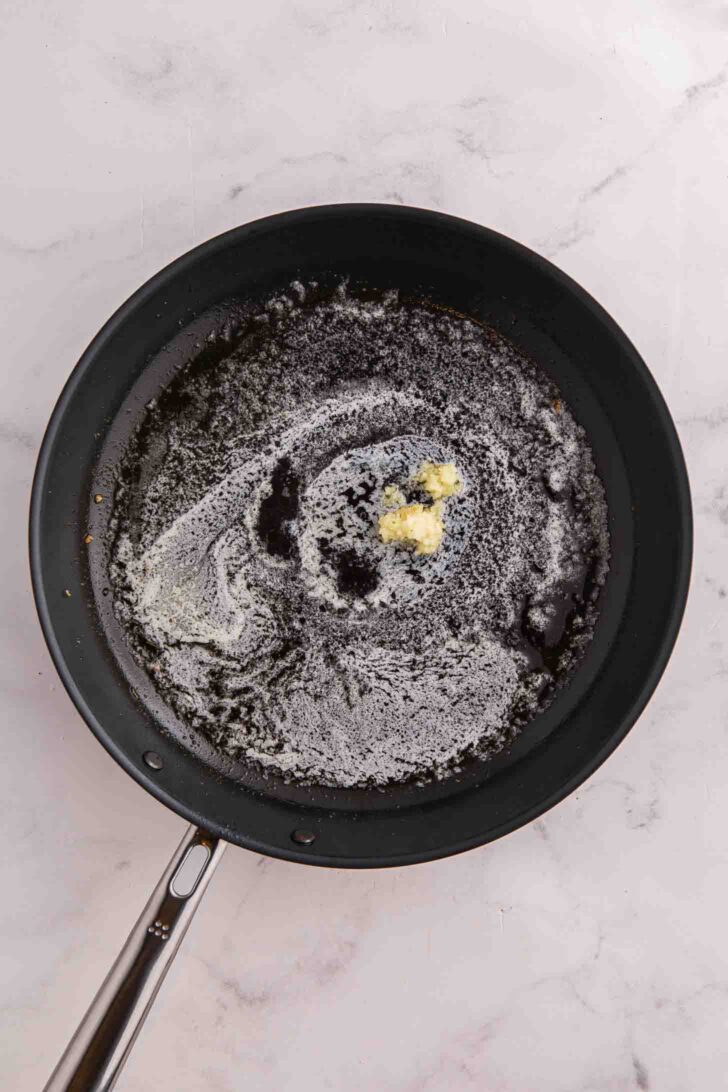 A black frying pan with melted butter and a small amount of minced garlic cooking on a marble countertop.