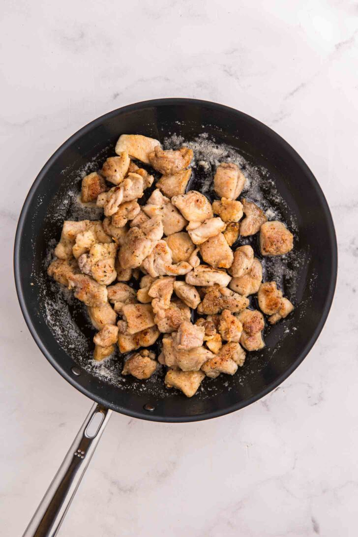 A black frying pan filled with cooked, seasoned pieces of chicken sits on a white marble surface.