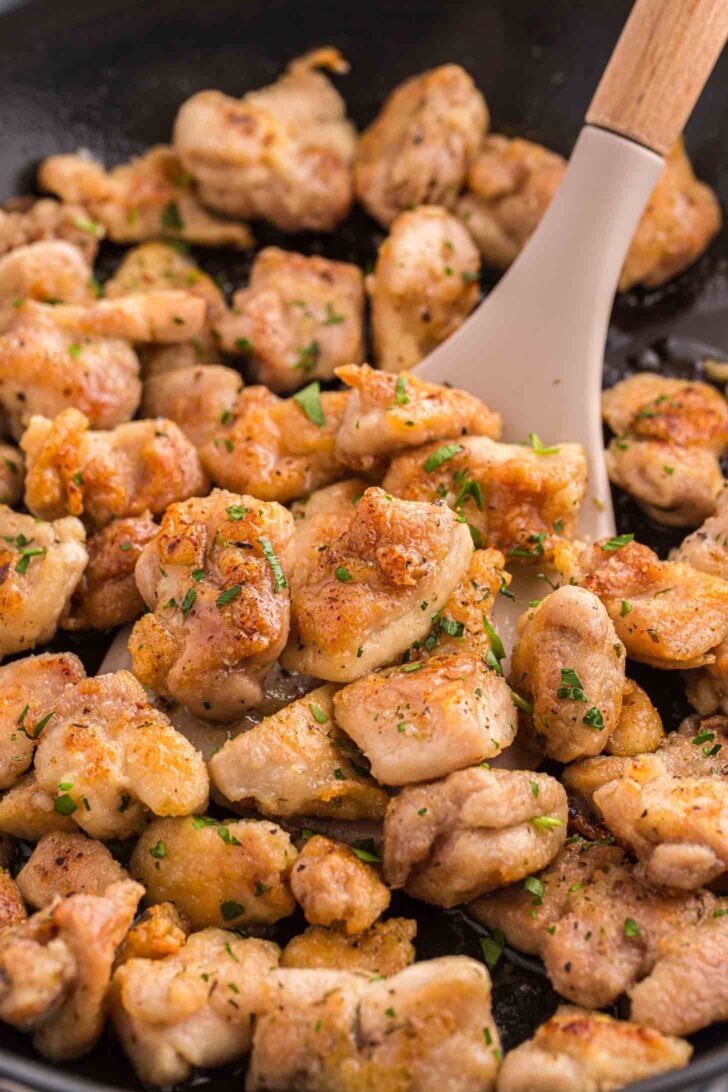 Close-up of golden-brown, seasoned chicken pieces being stirred in a skillet with a spatula. Some pieces are garnished with chopped herbs.