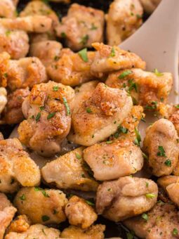 Golden brown, seasoned chicken pieces being sautéed in a pan with a spatula, garnished with chopped herbs.