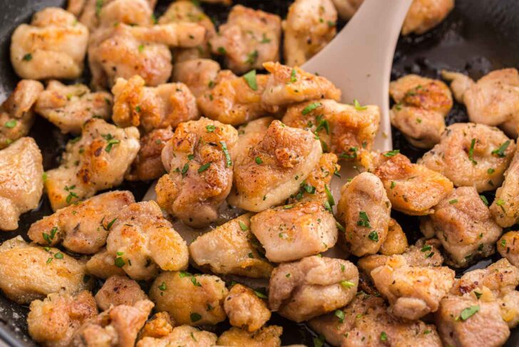 Golden brown, seasoned chicken pieces being sautéed in a pan with a spatula, garnished with chopped herbs.