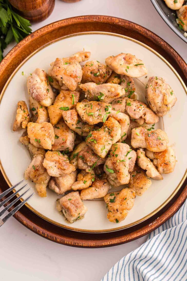 A plate of cooked, golden-brown chicken bites garnished with chopped herbs, set on a round wooden tray with a fork nearby.