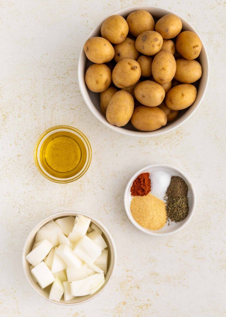 A bowl of small potatoes, a bowl of chopped onions, a small bowl with olive oil, and a small bowl containing salt, pepper, paprika, garlic powder, and dried herbs are arranged on a light-colored surface.