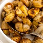 A close-up of roasted potatoes and onions in a white bowl, with a spoon scooping up some of the golden-brown, seasoned potato pieces.