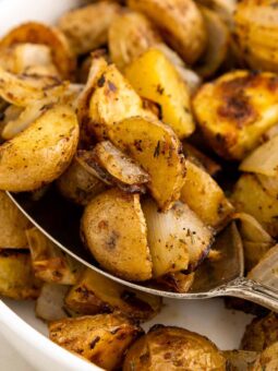 A close-up of roasted potatoes and onions in a white bowl, with a spoon scooping up some of the golden-brown, seasoned potato pieces.