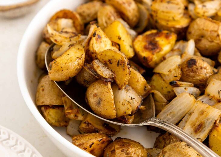A close-up of roasted potatoes and onions in a white bowl, with a spoon scooping up some of the golden-brown, seasoned potato pieces.