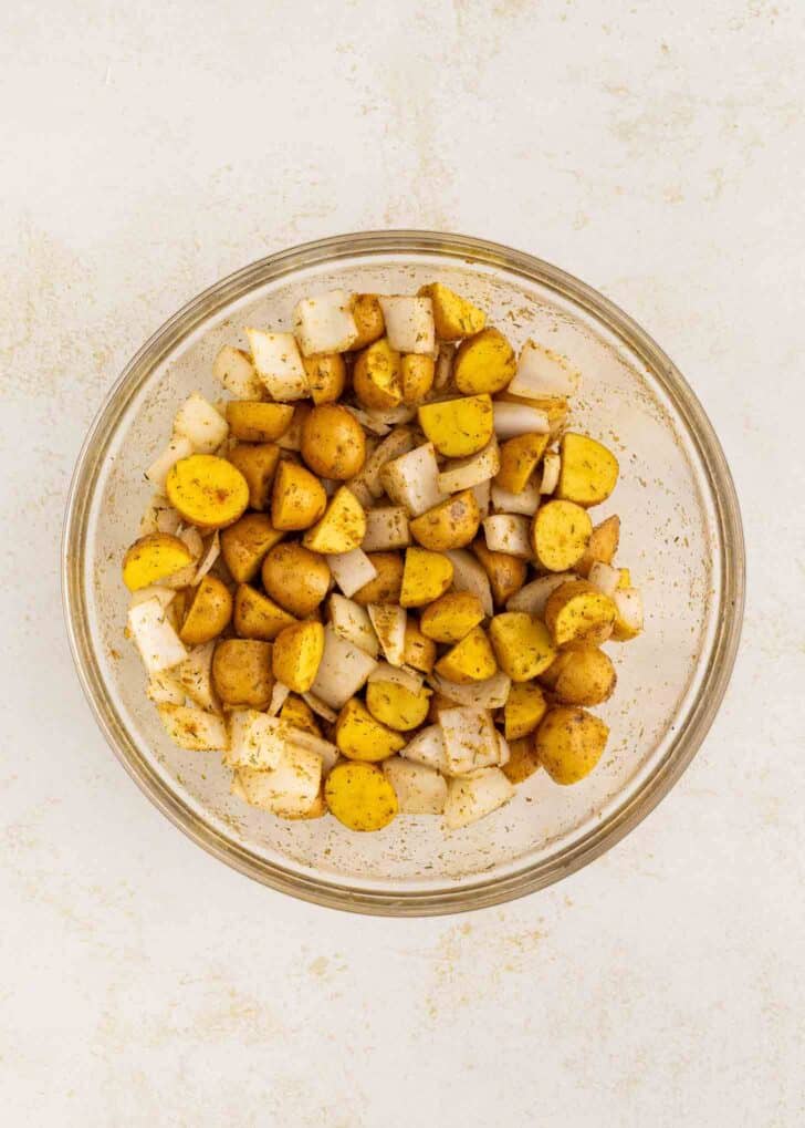 A clear glass bowl filled with diced yellow potatoes and white onions, coated with spices, sits on a light-colored surface.