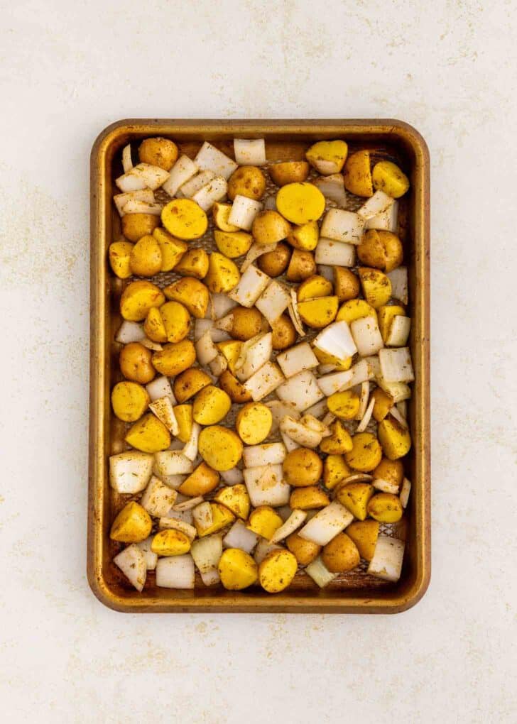 A baking sheet filled with chopped yellow potatoes and diced onions, seasoned and ready to roast, sits on a light-colored surface.