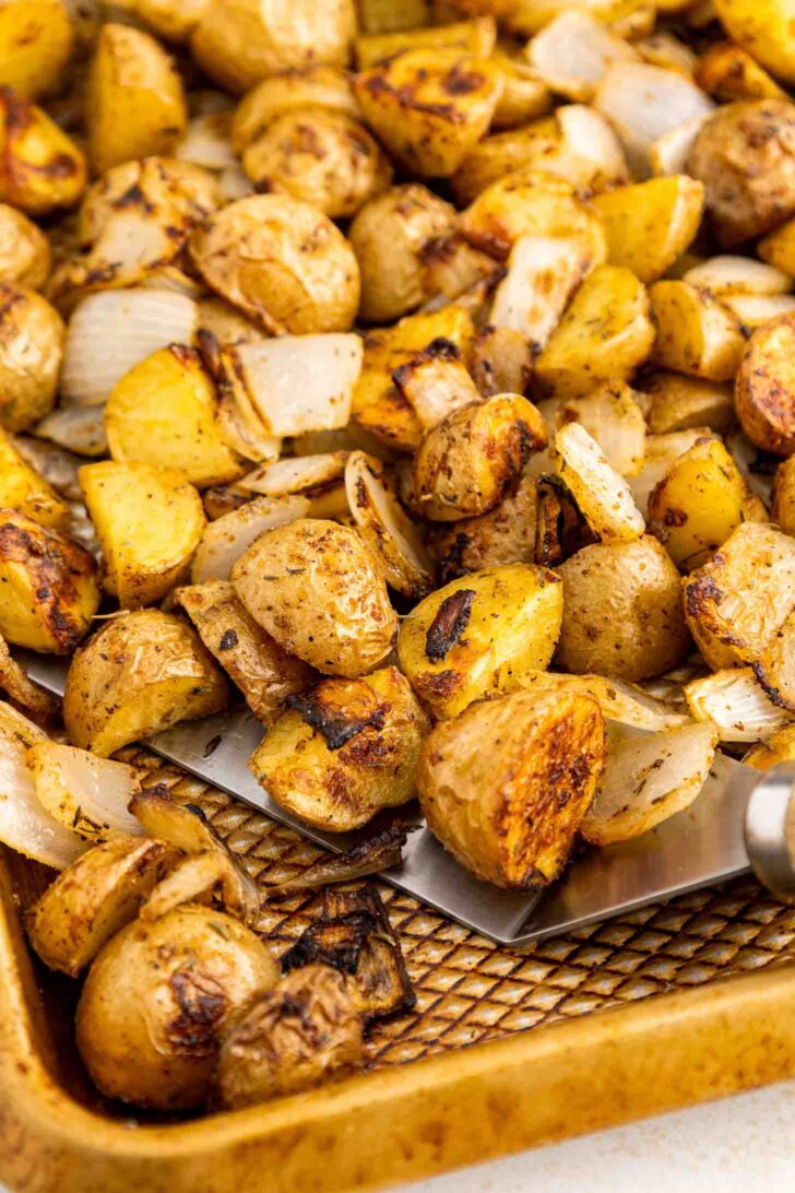 A baking sheet filled with roasted potato pieces and onions, seasoned and browned, with a metal spatula lifting some of the vegetables.
