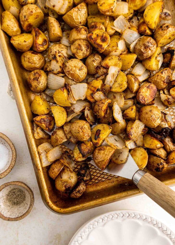 A baking sheet filled with roasted baby potatoes and chopped onions, seasoned and golden brown, with a spatula lifting a portion. Plates and small bowls are nearby.