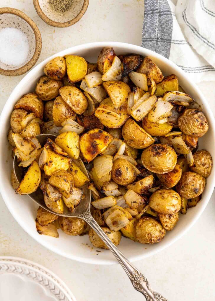 A white bowl filled with roasted potatoes and onions, seasoned with herbs, sits on a light surface with a serving spoon. Salt, pepper, and a striped napkin are nearby.