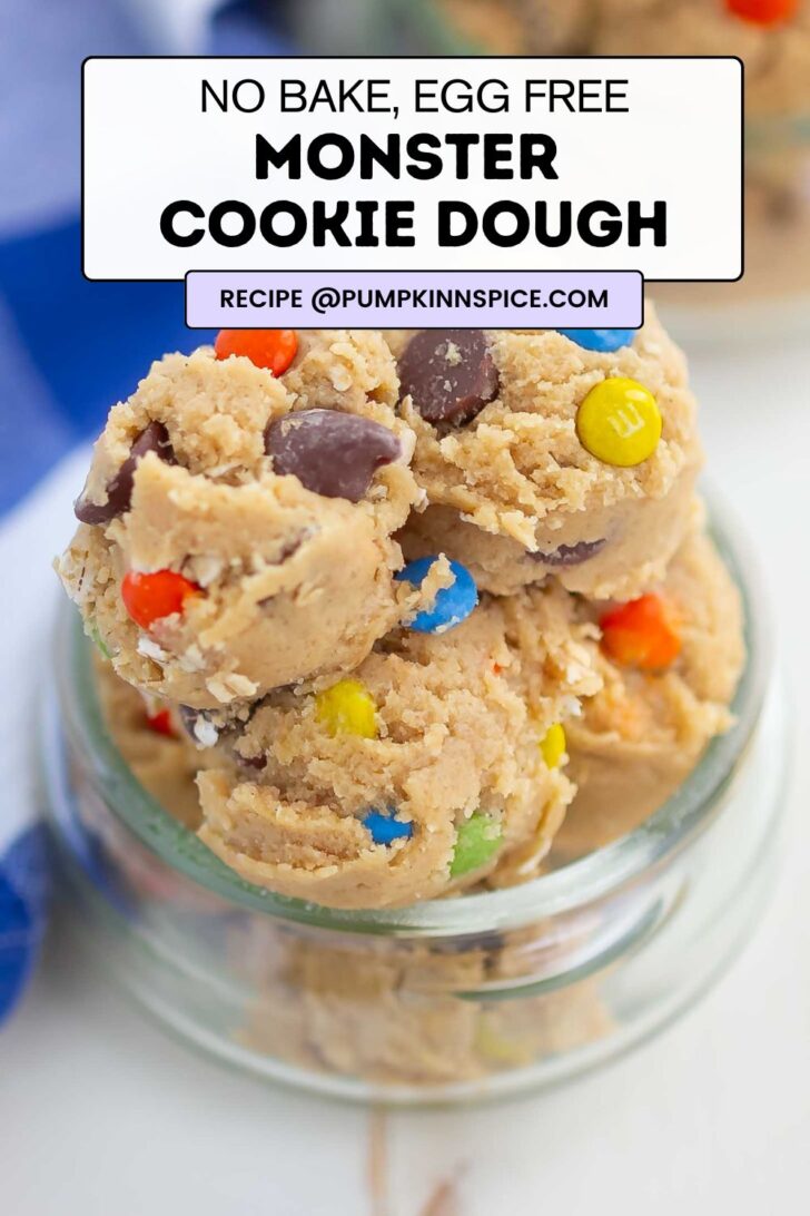 A jar filled with no-bake, egg-free monster cookie dough topped with colorful candy-coated chocolates and chocolate chips. A label on top shows the recipe title and source.