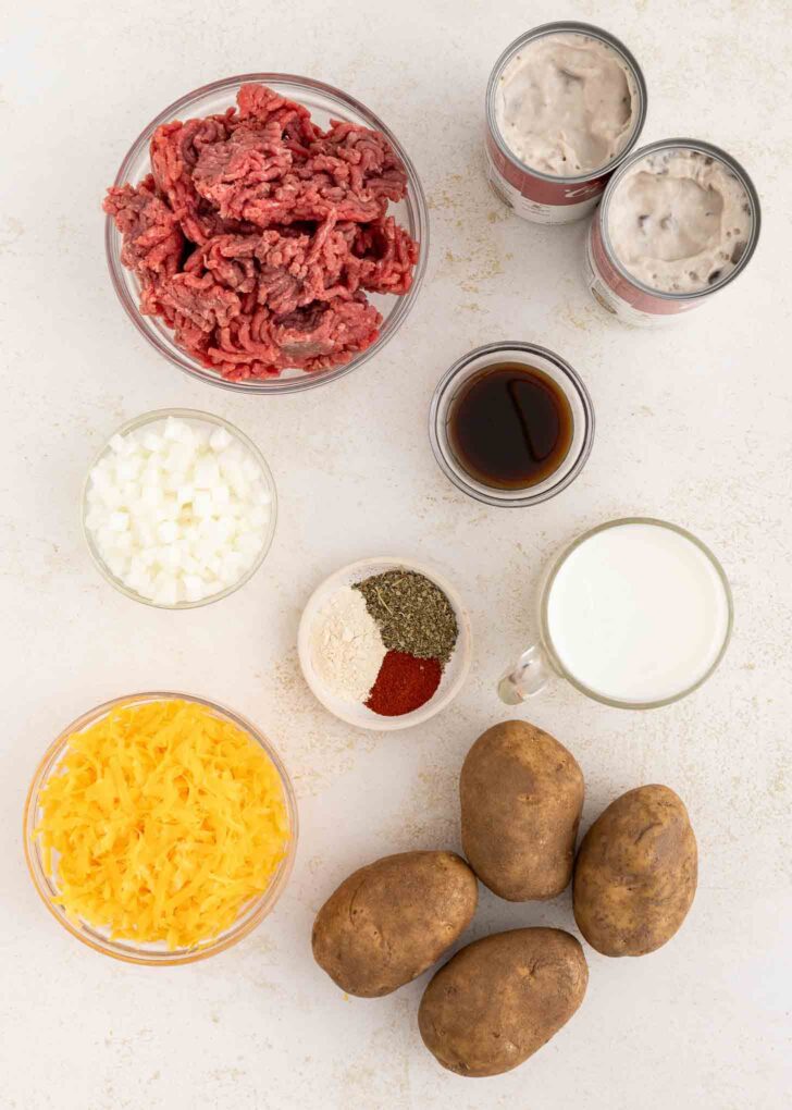 Ingredients laid out on a white surface: ground beef, canned soup, diced onions, shredded cheddar cheese, milk, Worcestershire sauce, seasonings, and four whole russet potatoes.