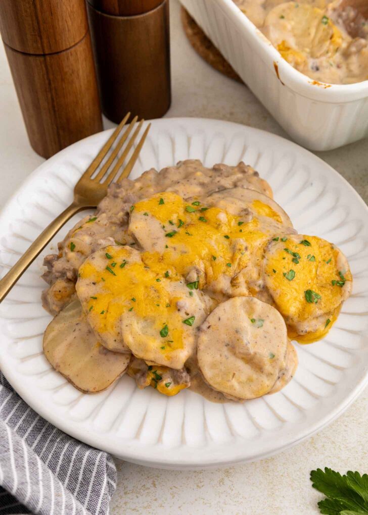 A white plate with cheesy hamburger potato casserole, featuring sliced potatoes, ground beef, creamy sauce, and melted cheese, garnished with parsley. A fork is on the plate.
