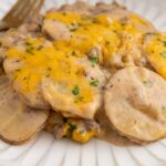 A serving of cheesy scalloped potatoes with ground beef, topped with melted cheese and chopped herbs, sits on a white plate with a gold fork beside it.