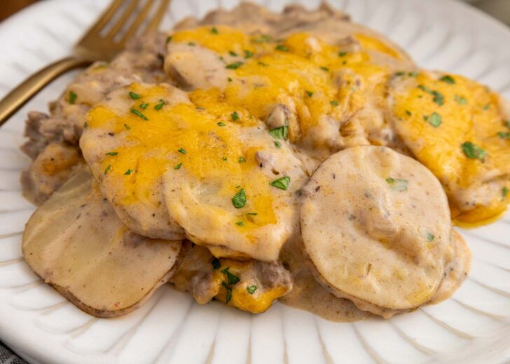 A serving of cheesy scalloped potatoes with ground beef, topped with melted cheese and chopped herbs, sits on a white plate with a gold fork beside it.