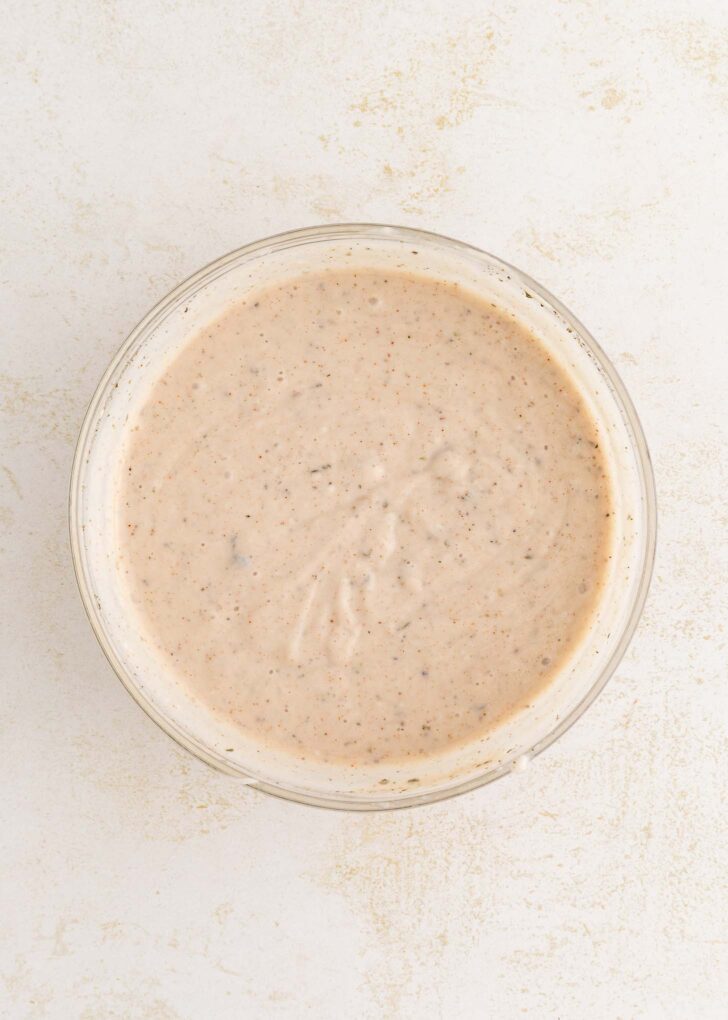 A glass bowl filled with creamy, speckled sauce sits on a light, textured surface.