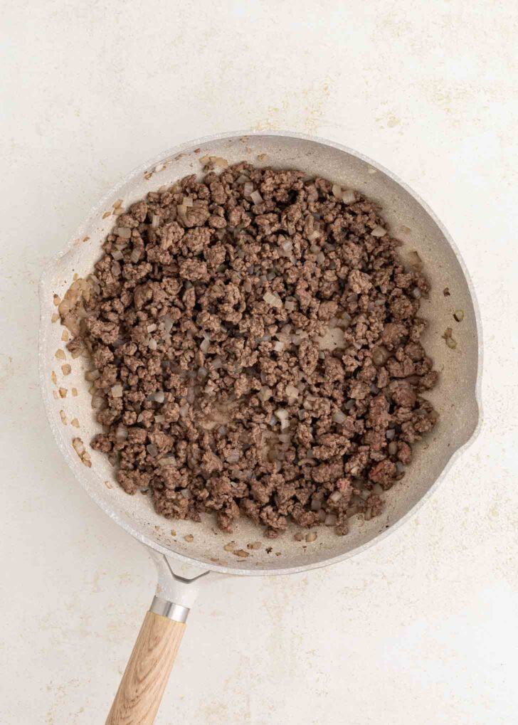 A frying pan with browned ground beef and diced onions on a light-colored surface.