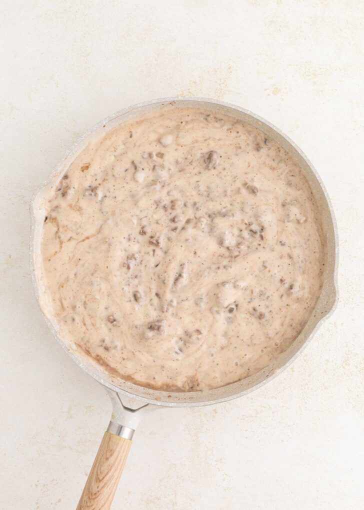 A skillet filled with creamy ground beef gravy, featuring visible chunks of sausage, sits on a light-colored surface.