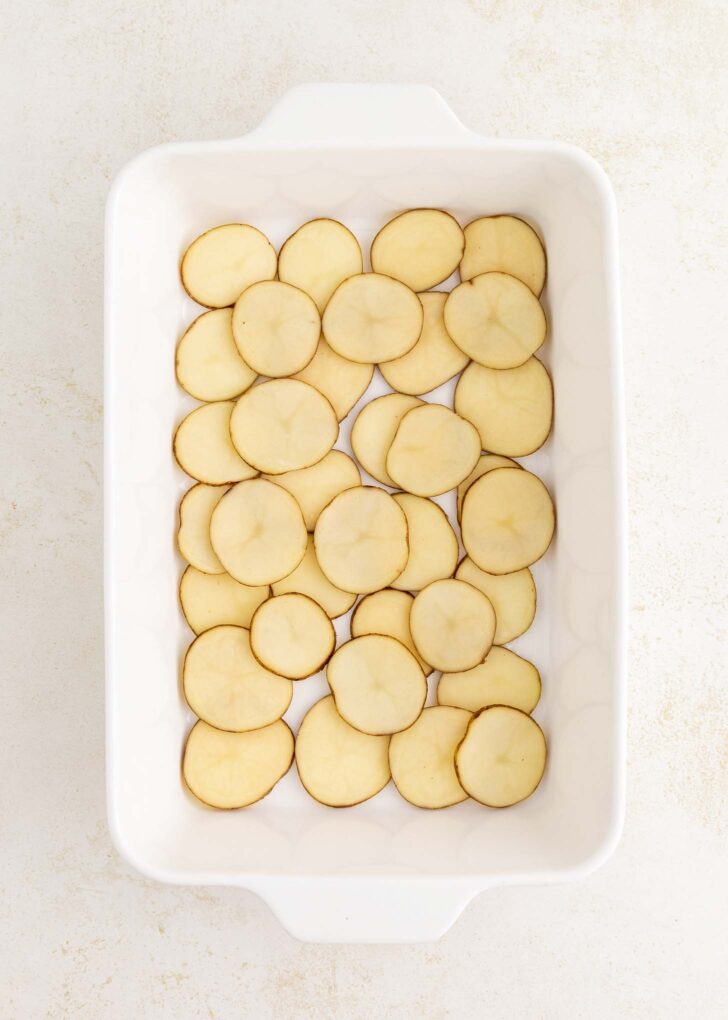 A white rectangular baking dish filled with a single layer of thinly sliced raw potatoes arranged neatly on the bottom.
