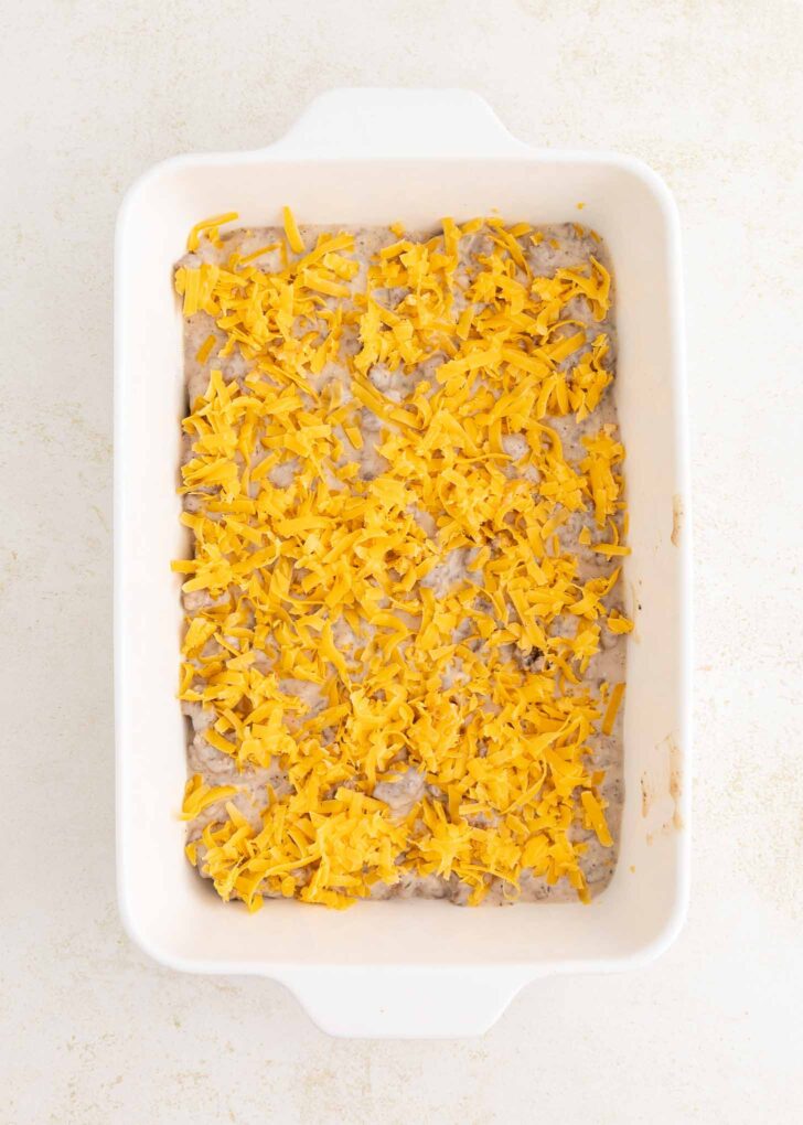 A white baking dish filled with a layer of creamy mixture topped with shredded cheddar cheese, ready to be baked. The dish is placed on a light-colored surface.