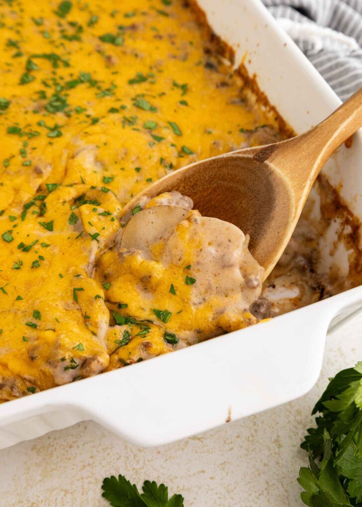 A wooden spoon scoops a serving of cheesy casserole topped with melted cheddar and chopped parsley from a white baking dish.