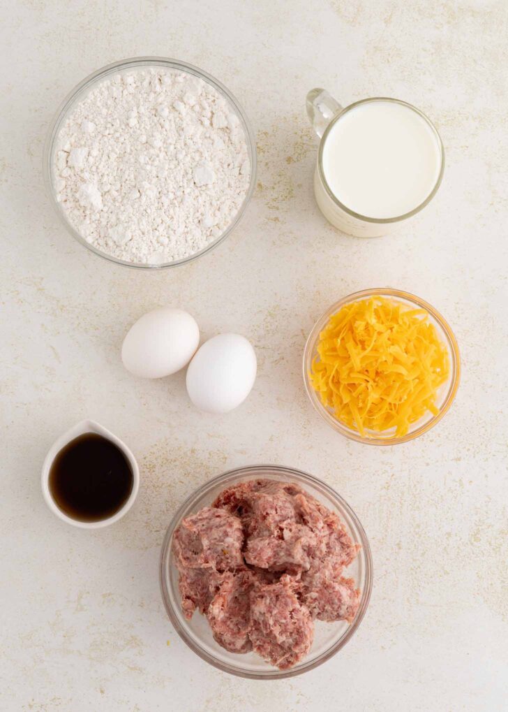 Top-down view of baking ingredients: a bowl of flour, a glass of milk, two eggs, shredded cheese, a small cup of vanilla extract, and a bowl of ground sausage arranged on a light surface.