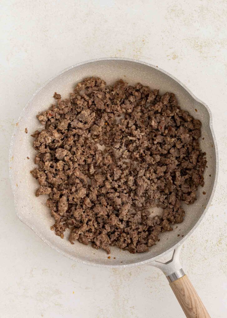 A frying pan with cooked, browned sausage sits on a light-colored surface. The beef is crumbled and evenly spread across the pan.