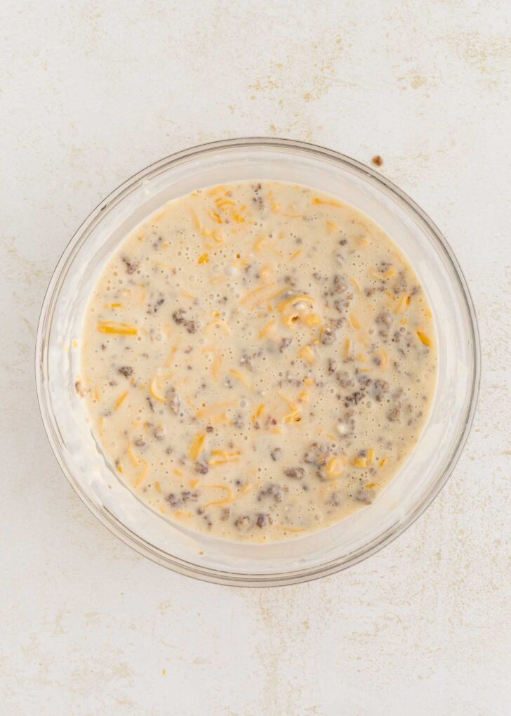 A glass bowl filled with a mixture of ground beef, shredded cheese, pancake mix, and beaten eggs, sitting on a light-colored surface.