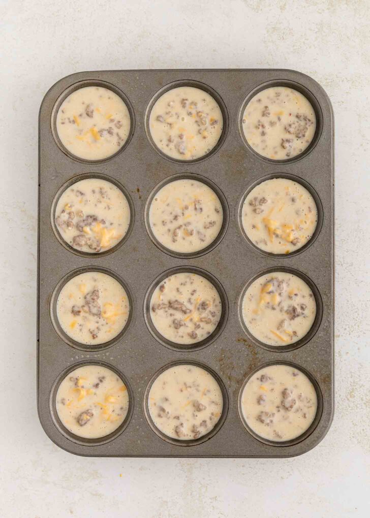 A 12-cup muffin tin filled with unbaked muffin batter containing visible sausage crumbles and shredded cheese, set on a light-colored surface.