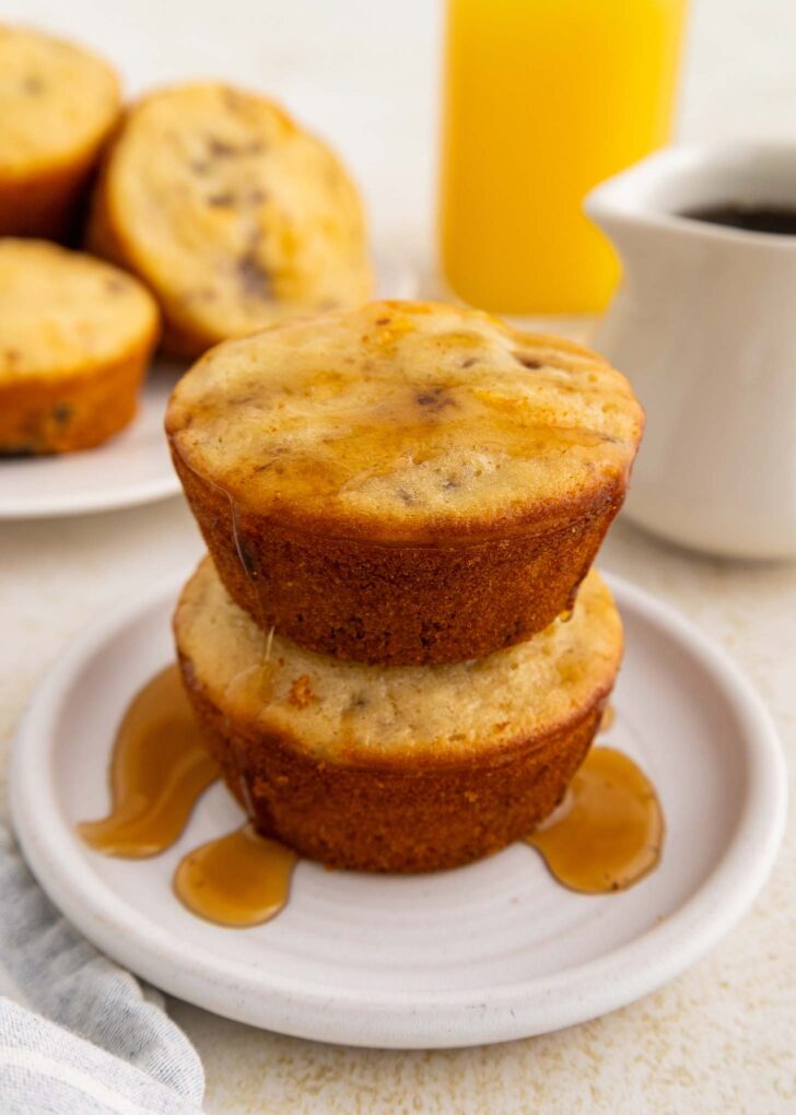 Two golden muffin-like pancakes stacked on a white plate, drizzled with syrup. In the background are more pancake muffins on a plate, a glass of orange juice, and a white cup of coffee.