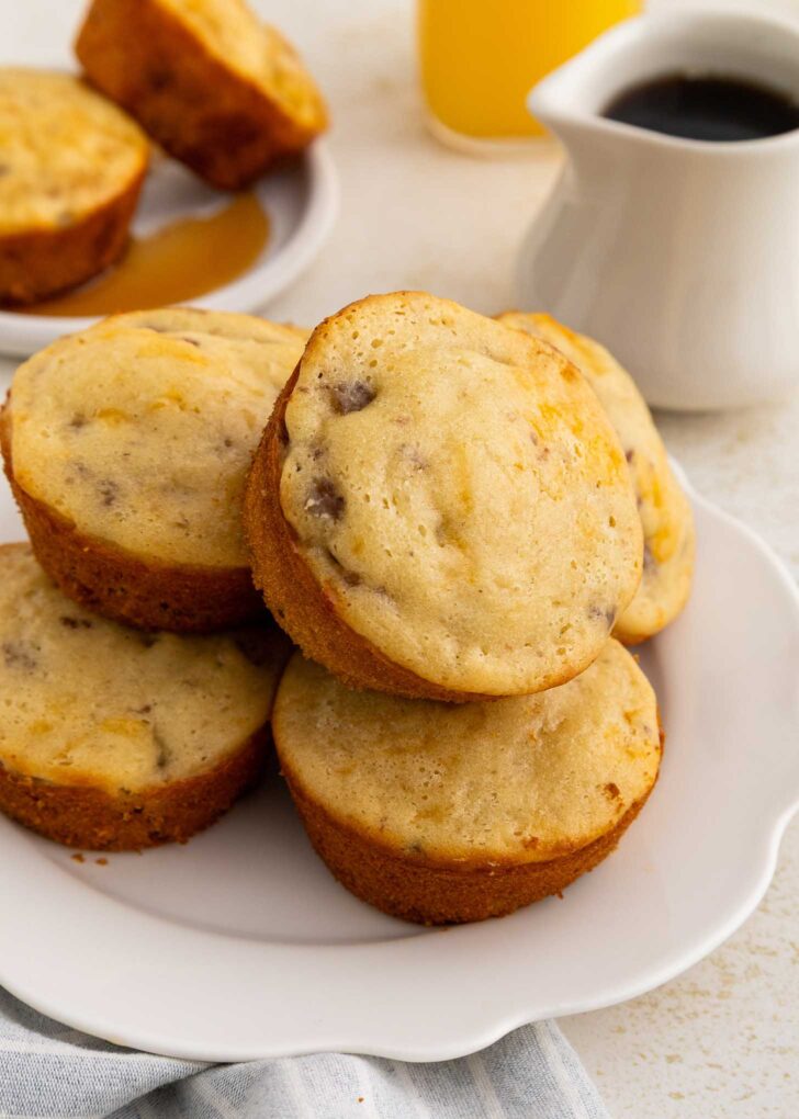 A plate of golden-brown muffins with visible sausage sits on a white plate. In the background are a glass of orange juice, a white pitcher, and more muffins on a small plate.