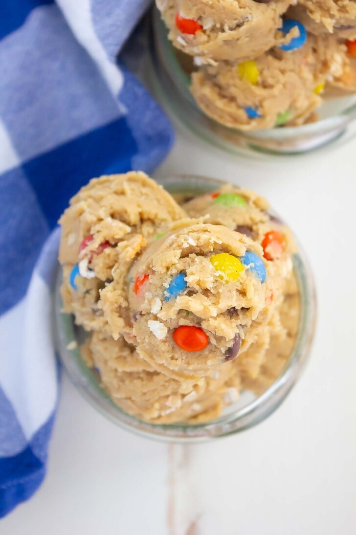 Scoops of edible cookie dough with colorful candy-coated chocolates in glass jars, next to a blue-and-white checkered cloth, on a white surface.