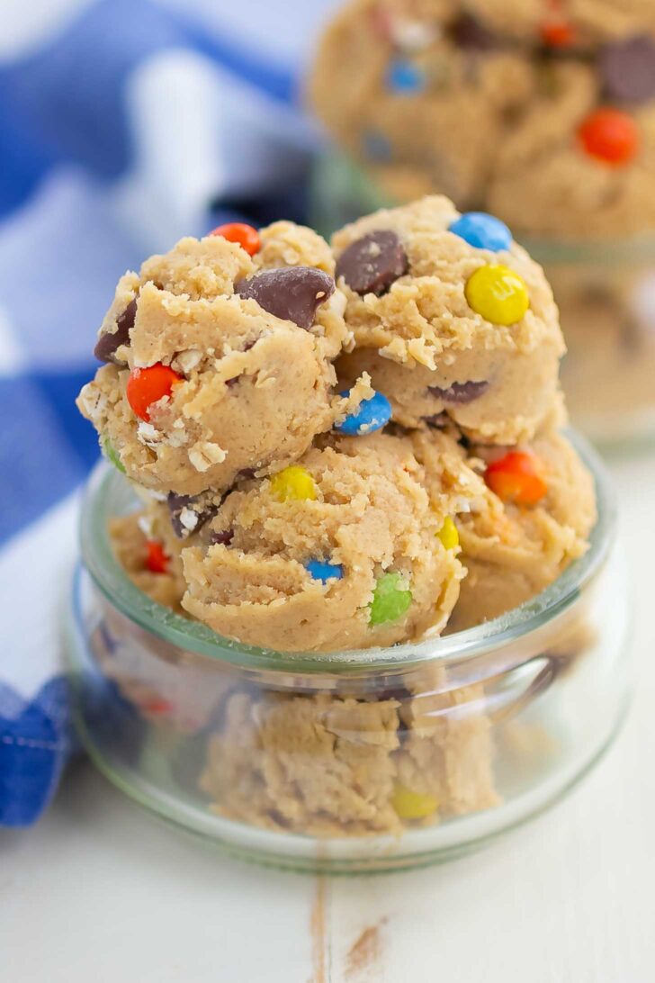 Scoops of cookie dough with colorful candy-coated chocolates and chocolate chips stacked in a small glass jar, with more dough blurred in the background.