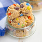 Scoops of cookie dough with colorful candy-coated chocolates and chocolate chips in a small glass jar, next to a blue and white checkered cloth.